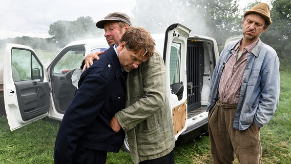 Szenenbild aus der 80. Büttenwarder-Folge "Zwei Steinadler": Hajo Narkmeyer hält den benommenen Fahrer des verunglückten Goldtransporters im Arm, Adsche beäugt das skeptisch. © NDR/Uwe Ernst Foto: Uwe Ernst