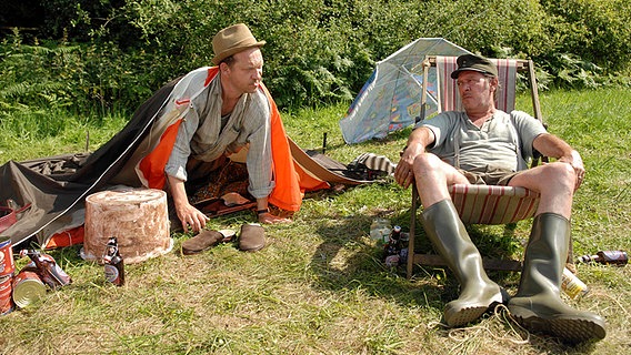 Szenenbild aus der 26. Büttenwarder-Folge "Endlich Urlaub": Adsche baut das Zelt auf, Brakelmann hat es sich im Liegestuhl gemütlich gemacht und schaut ihm zu. © NDR/Nicolas Maack 