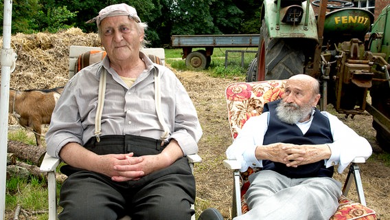 Szenenbild aus der 18. Büttenwarder-Folge "Pflegeparadies": Onkel Krischan und Herr Buschbohn sitzen im Gartenstuhl vor einem Misthaufen. © NDR/Sandra Hoever 