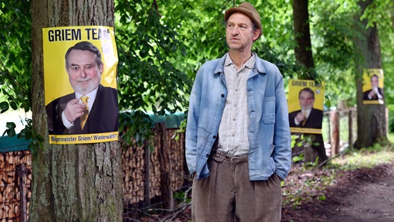 Szene aus der 96. Büttenwarder-Folge "Nur noch 14 Tage": An einem Baum hängt ein Wahlplakat, ein Mann blick darauf. © NDR/Nicolas Maack 