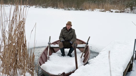 Szenenbild aus der 52. Büttenwarder-Folge "Patchwork": Adsche sitzt im Boot auf dem eingeschneiten Dorfteich. © NDR/Nico Maack Foto: Nico Maack