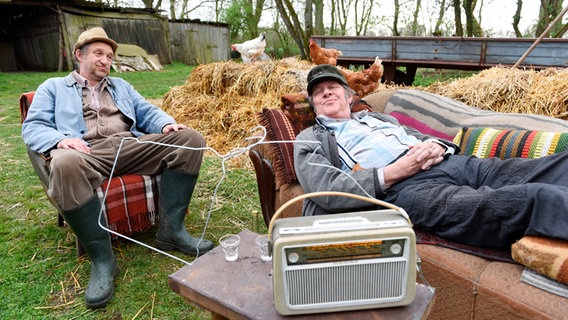 Szenenbild aus der 64. Büttenwarder-Folge "Radio": Adsche und Brakelmann sitzen draußen und hören Radio. © NDR/Nicolas Maack Foto: Nicolas Maack
