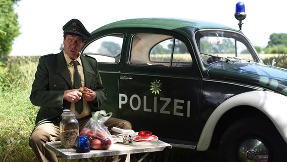 Szenenbild aus der 66. Büttenwarder-Folge "Rekord": Polizist Peter macht Picknick am Wegesrand und beobachtet Leute. © NDR/Nicolas Maack Foto: Nicolas Maack