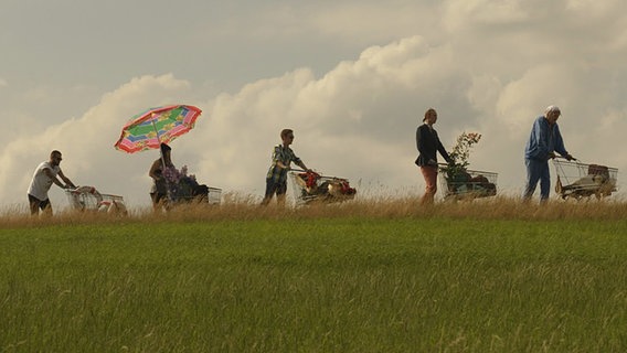 Onkel Krischan führt die Meute junger Männer mit Einkaufswagen über eine Weide an. © NDR/Nicolas Maack Foto: Nicolas Maack