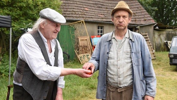 Szenenbild aus der 65. Büttenwarder-Folge "Rosenkrieg": Ein alter Mann (Onkel Krischan) gibt einem anderen Mann (Adsche) eine Tomate. © NDR/Nicolas Maack Foto: Nicolas Maack