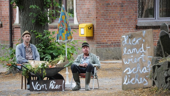 Szenenbild aus der 55. Büttenwarder-Folge "Stau": Brakelmann und Adsche verkaufen Gemüse an der Straße beim Dorfkrug. Sie sitzen auf ihren Gartenstühlen mit dem Sonnenschirm. © NDR/Nico Maack Foto: Nico Maack