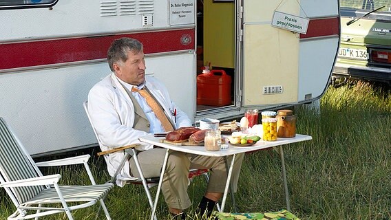 Dr. Rufus Kloppstedt (Veit Stübner) hat seinen Wohnwagen auf der großen Dorfwiese abgestellt. Er ist mit sich und der Welt zufrieden und genießt sein Frühstück. © NDR/Sandra Hoever 