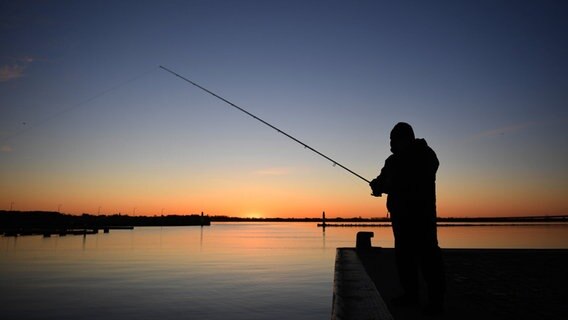 Stralsund: Die aufgehende Sonne vor einem Angler taucht den Himmel im Hafen in ein warmes Licht © NDR Foto: Stefan Sauer/dpa-Zentralbild/dpa