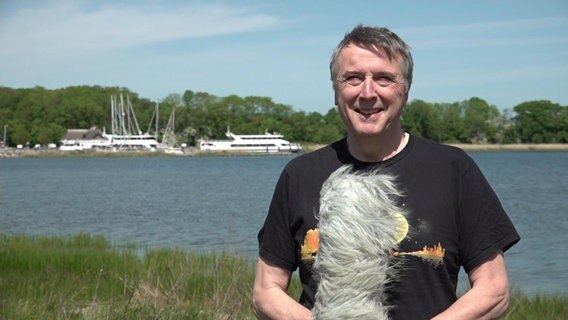 Uwe Ulbrich steht mit einem Mikrofon vor einem Gewässer. © NDR 