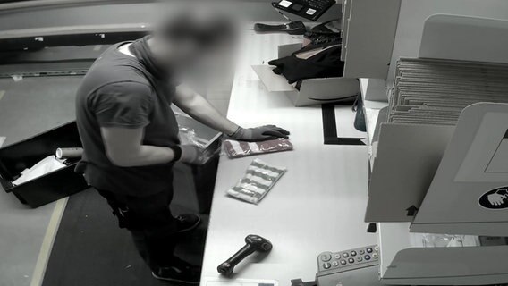 Eine unkenntlich gemachte Person steht vor einem Tisch und bearbeitet einen vor ihm liegenden Gegenstand. Eine unkenntlich gemachte Person steht vor einem Tisch und bearbeitet einen vor ihm liegenden Gegenstand. © NDR/ARD Foto: Screenshot