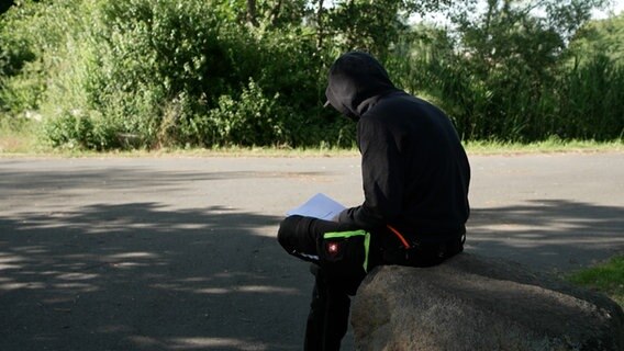 Person mit Kapuze sitzt und hält Papier in der Hand © NDR 