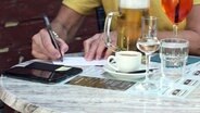 Person füllt einen Zettel aus, auf dem Tisch stehen Kaffee und Bier. © NDR Foto: Screenshot