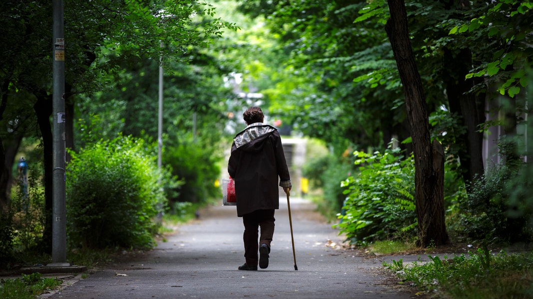 Eine ältere Frau geht auf einen Stock gestützt einen Weg entlang.
