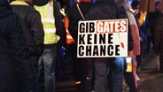 Thumbnail: Eine Person mit einem Schild auf dem Rücken, auf dem steht: Gib Gates keine Chance. © NDR Foto: Screenshot