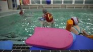 Kinder und Erwachsene schwimmen in einem Becken in einer Schwimmhalle, im Vordergrund Schwimmhilfen. © NDR Foto: Screenshot