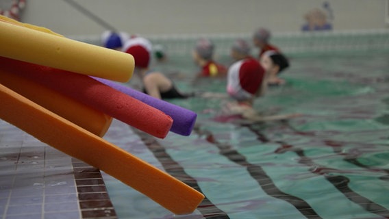 Kinder und Erwachsene schwimmen in einem Becken in einer Schwimmhalle, im Vordergrund Schwimmhilfen. © NDR Foto: Screenshot