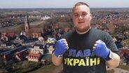 Thumbnail: Ein Mann mit Boxhandschuhen und einem ukrainischen T-Shirt, im Hintergrund die niedersäschsische Gemeinde Molbergen. © NDR Foto: screenshot