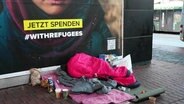 Ein Schlafsack liegt auf dem Boden vor einem Plakat, Symbolbild Obdachlosigkeit © Screenshot 
