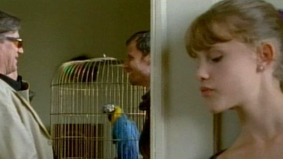 Lilly steht hinter neben einer Tür und belauscht zwei Männer an einem Papageienkäfig. © NDR 
