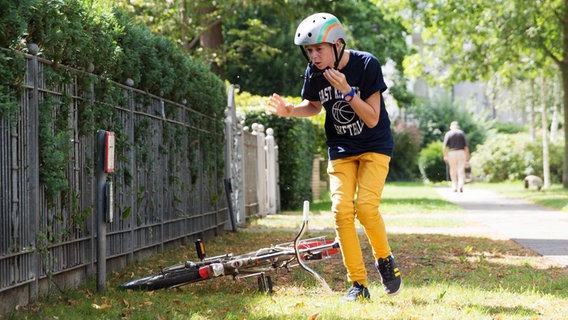 Niklas (Jaden Dreier) steht geduckt und öffnet seinen Helmgurt. Neben ihm liegt sein Klapprad. © NDR Foto: Claudia Timmann