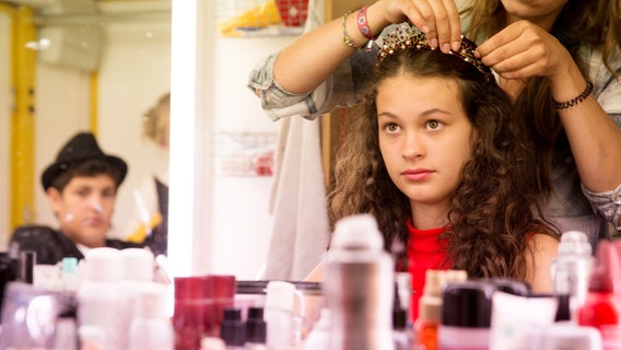 Ceyda (Merle de Villiers) sitzt vor dem Spiegel in der Maske und bekommt von der Visagistin ein Diadem ins Haar gesteckt. © NDR Foto: Claudia Timmann