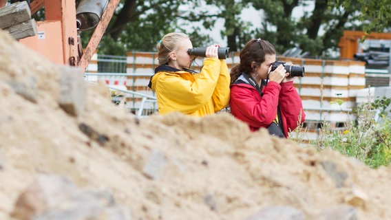 Jessi (Martha Fries) und Ceyda (Merle de Villiers) stehen hinter einem Sandhügel und schauen durch ein Fernglas und eine Kamera. © NDR Foto: Claudia Timmann