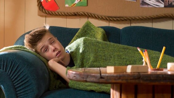 Niklas (Jaden Dreier) liegt zugedeckt auf dem Sofa und öffnet ein Auge. © NDR Foto: Claudia Timmann