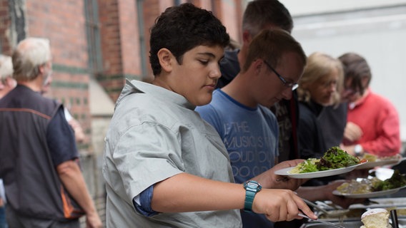 Danilo Kamber steht mit einem umgedrehten Hemd bekleidet am Buffet mit einem Teller Salat in der Hand. © NDR Foto: Claudia Timmann