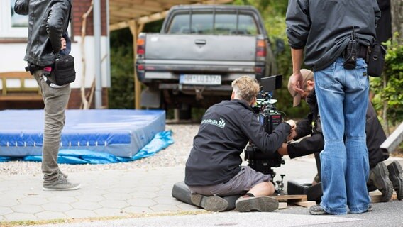 Der Kameramann und sein Assistent knien auf dem Boden neben einer Kamera. Im Hintergrund sieht man eine blaue Matte und einen Pick Up. © NDR Foto: Claudia Timmann