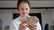 Leon (Luka Krause) hält lächelnd Geldscheine in der Hand. © NDR/Studio HH Foto: Romano Ruhnau