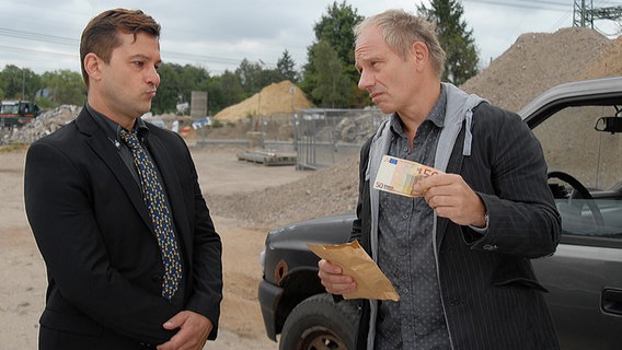 Harald Stucke (rechts, Michael Lott) steht vor einem Mann und hält einen 50 Euro Schein in der Hand. © NDR/Studio HH Foto: Romano Ruhnau