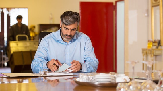 Navid (Neil Malik Abdullah) sitzt an einem Schreibtisch im Hintergrund erscheint ein Bote mit Kisten auf einer Sackkarre. © NDR Foto: Claudia Timmann