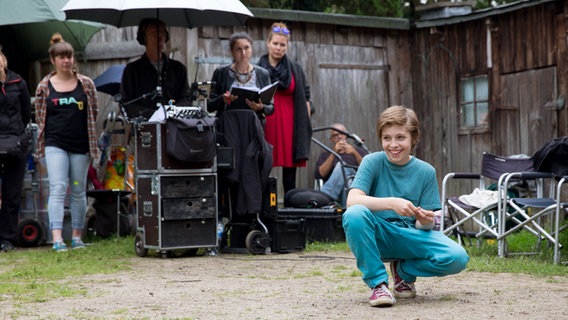 Till (Otto von Grevenmoor) hockt lächelnd auf dem Boden, vor dem Schuppen. Hinter ihm stehen Dreharbeiter. © NDR Foto: Claudia Timmann