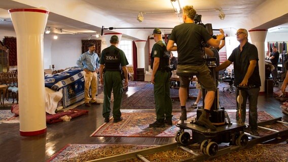 Navid (Neil Malik Abdullah) steht im Teppichkontor vor zwei Zollbeamten. Drum herum steht das Kamerateam. © NDR Foto: Claudia Timmann