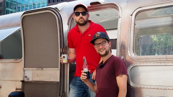 Zwei Teammitglieder stehen mit Getränken in der Hand vor einem silbernen Wohnwagen. © NDR Foto: Claudia Timmann