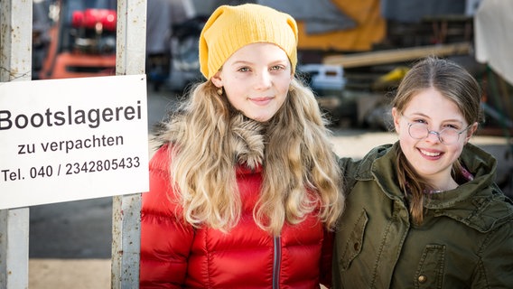 Stella (Zoë Malia Moon) und Pinja (Sina Michel) stehen neben einem Schild der Bootslagerei. © NDR/StudioHH Foto: Boris Laewen