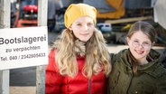 Stella (Zoë Malia Moon) und Pinja (Sina Michel) stehen neben einem Schild der Bootslagerei. © NDR/StudioHH Foto: Boris Laewen