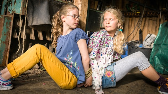 Pinja (Sina Michel) und Stella (Zoë Malia Moon) sitzen mit gefesselten Händen Rücken an Rücken. © NDR/Studio HH Foto: Boris Laewen