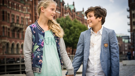 Stella (Zoë Malia Moon, li.) und Ramin (Jann Piet, re.) stehen Hand in Hand vor der Speicherstadt. © NDR/Studio HH Foto: Boris Laewen