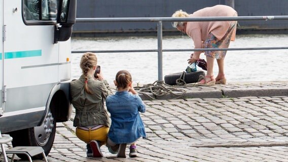 Pinja (Sina Michel) und Jale (Ava Sophie Richter) hocken versteckt neben einem Wohnmobil und richten ihre Handys auf die Trickbetrügerin, die sich über ein Netz mit Brieftaschen beugt. © NDR Foto: Claudia Timmann