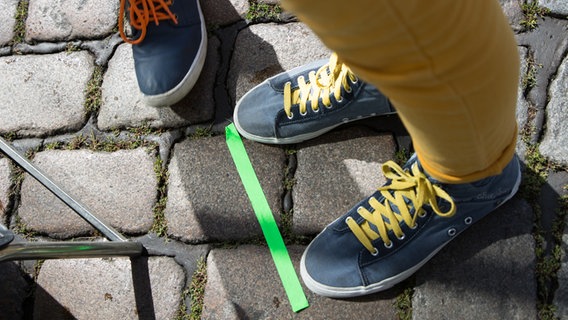 Ein grüner Klebestreifen klebt auf dem Kopfsteinpflaster, dahinter sieht man Schuhe. © NDR Foto: Claudia Timmann