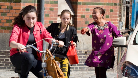 Sara (Antonia Büchel) fährt auf dem Rad an Oma Dschami (Lilay Huser) vorbei und hält eine rote Handtasche in der Hand. © NDR Foto: Claudia Timmann