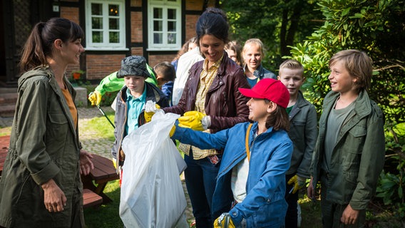 Jale (Ava Sophie Richter, 3.vr) zeigt Eva Dreyer (Anne Weber, links) und Frau Schultze (Katinka Auberger, Mitte) stolz einen Müllbeutel, drumherum stehen weitere Kinder. © NDR/Studio HH Foto: Boris Laewen