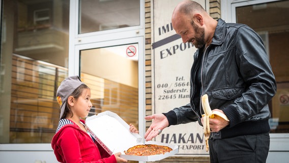 Jale (Ava Sophie Richter) hält Fenno (Alexander Grünberg) einen geöffneten Pizzakarton hin. © NDR/Studio HH Foto: Boris Laewen