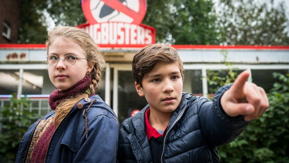 Pinja (Sina Michel) und Ramin (Jann Piet) stehen vor einem Schild mit der Aufschrift “Bugbusters”. © NDR/Studio HH Foto: Boris Laewen