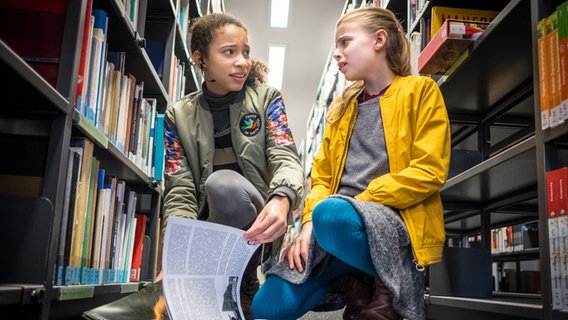 Lisha (Emma Roth) und Alice (Emilia Flint) hocken vor Bücherregalen und blättern in einem Buch. © NDR/Studio HH Foto: Boris Laewen