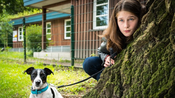 Mia Goldman (Marleen Quentin) hält einen Hund an der Leine und schaut hinter einem Baum hervor. © NDR Foto: Boris Laewen