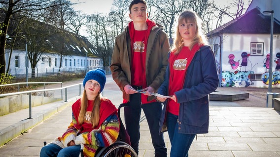 Pippa, Fritz und Paula tragen rote Pullover und verteilen Flyer auf dem Schulhof. © NDR/Letterbox Foto: Boris Laewen