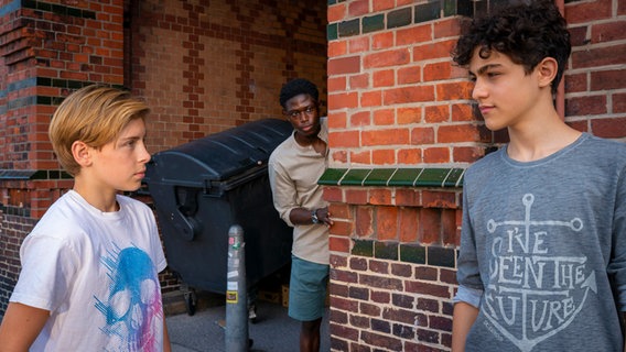 Jonny und Tarun schauen sich an, im Hintergrund schaut ein schwarzer Mann um die Ecke. © NDR/Letterbox Foto: Boris Laewen