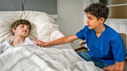 Tarun legt einem Jungen, der in einem Krankenhausbett liegt, eine Hand auf die Schulter. © NDR/Letterbox Foto: Boris Laewen